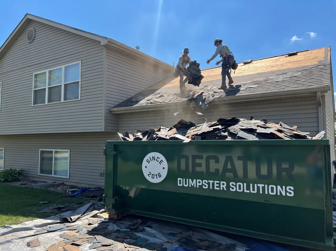 Green roll-off dumpster beside a house during a roof tear-off, filled with old shingles